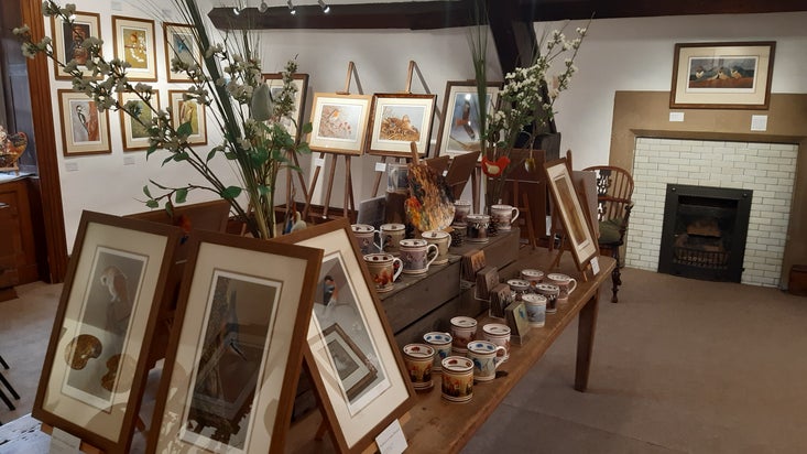 Framed artwork on the walls in a wooden-beamed room. There are framed works on easels and mug and flower displays on a table in the centre of the room.